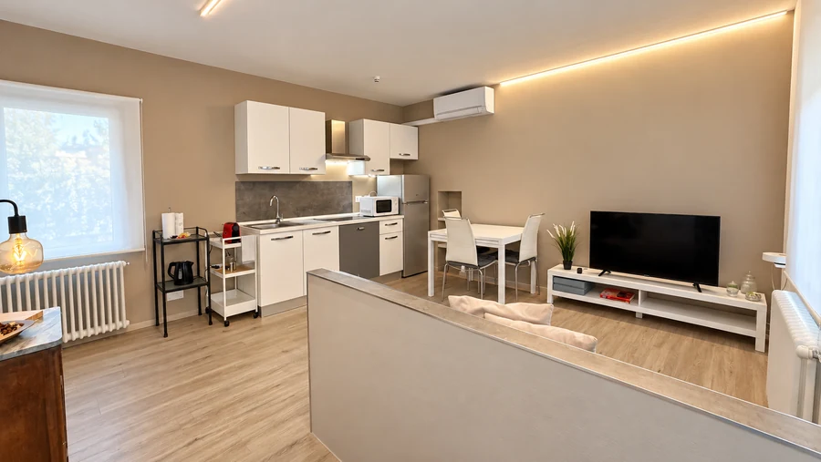 Zona giorno open space di Alkecè White con cucina attrezzata, tavolo da pranzo, TV e luce LED a soffitto